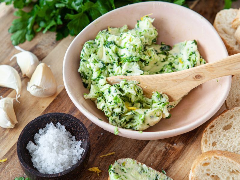 Kräuterbutter Rezept - Schale mit angerührter Kräuterbutter, daneben Knoblauch, Salz, Petersilie und Brot